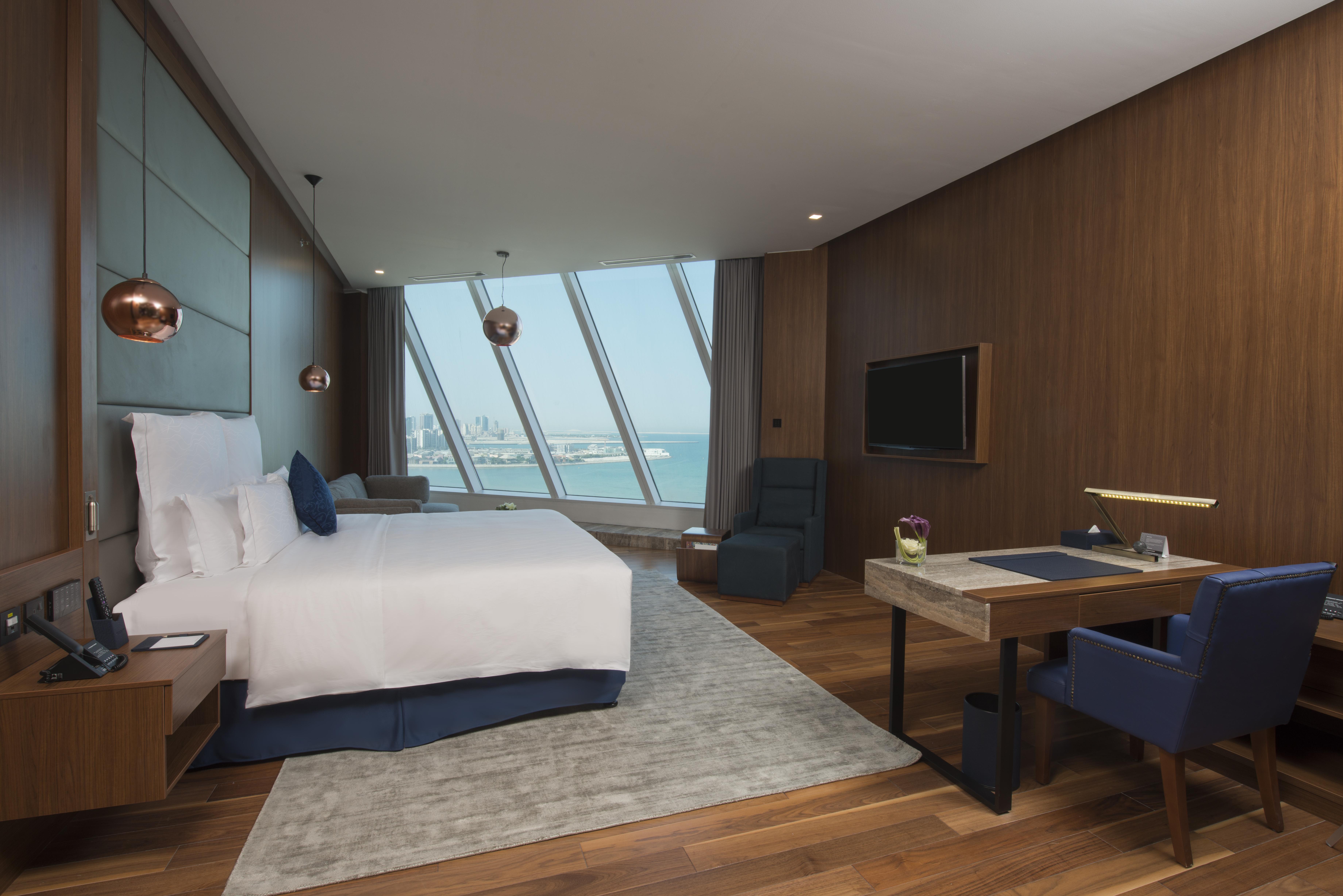 Wyndham Grand Manama Hotel Exterior photo The photo displays a modern hotel room featuring a large double bed with white linens and a blue bed skirt. The room has a stylish and contemporary design, characterized by wooden paneling and warm tones. Large windows offer a panoramic view of the o