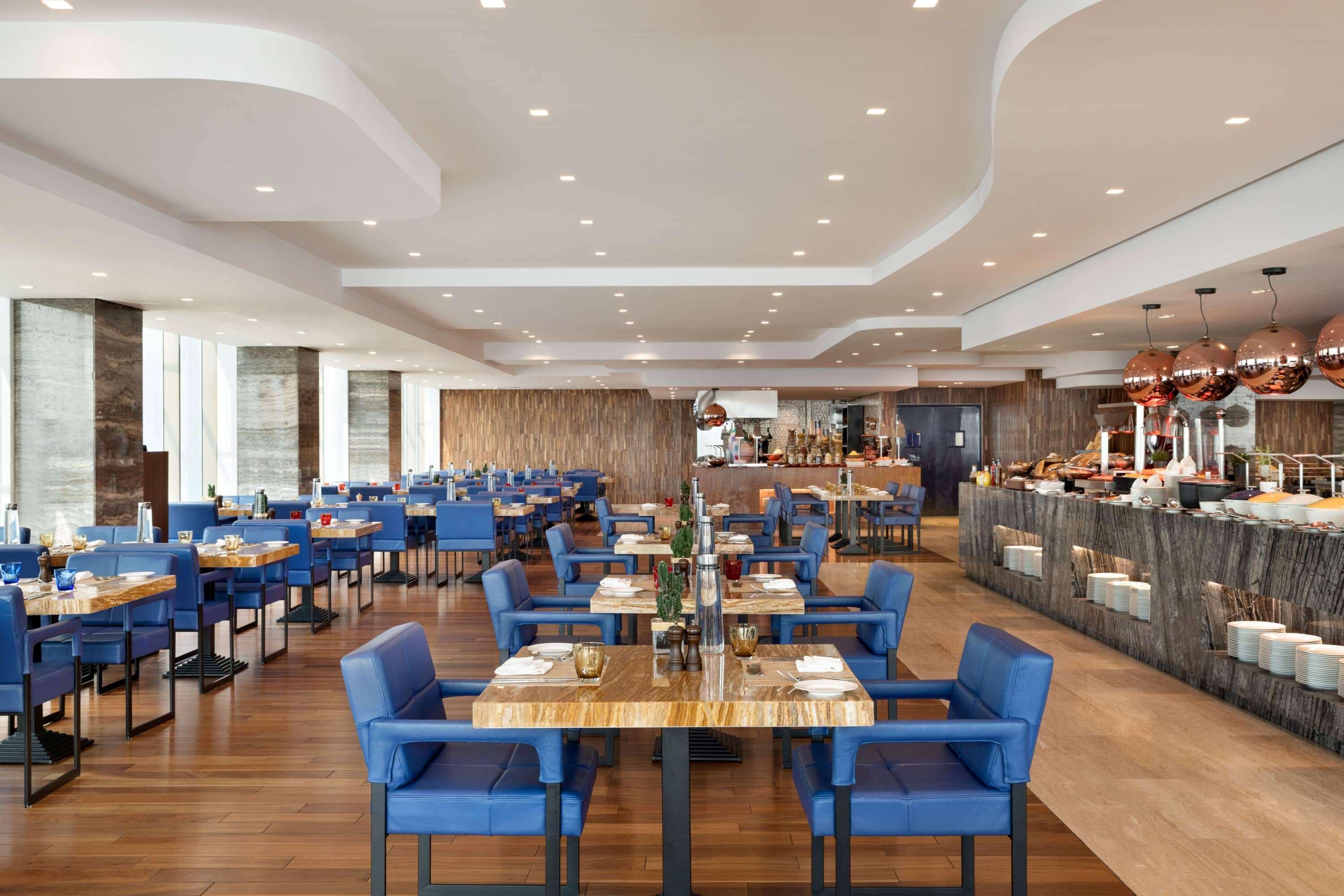Wyndham Grand Manama Hotel Exterior photo The image displays a modern restaurant interior. It features elegantly arranged tables with blue upholstered chairs. The flooring is made of wood, contributing a warm ambiance. The ceiling has a contemporary design with ample lighting that brightens 