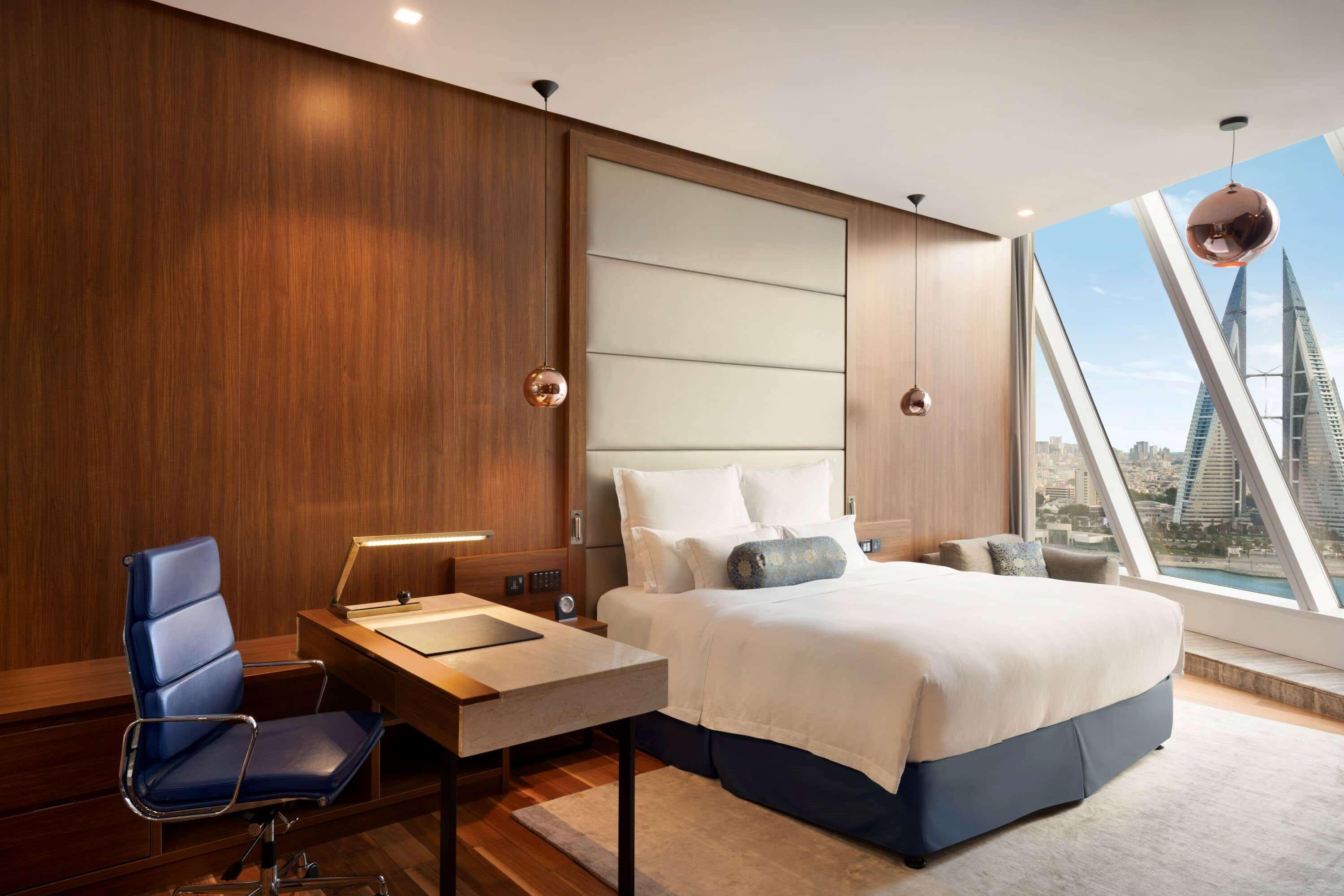 Wyndham Grand Manama Hotel Exterior photo The photo shows a modern hotel room featuring a large bed with white linens and a decorative pillow. There is a wooden accent wall behind the bed, adding warmth to the space. A stylish desk with a blue office chair is positioned next to the bed, and 