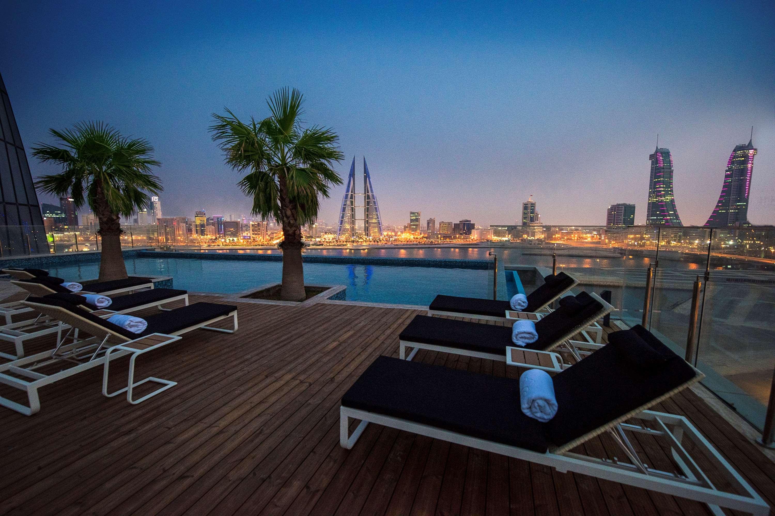 Wyndham Grand Manama Hotel Exterior photo The photo features a rooftop setting with several lounge chairs arranged alongside a pool. The chairs are white with dark cushions and have rolled towels placed on them. In the background, there are tall buildings illuminated by city lights, with a s