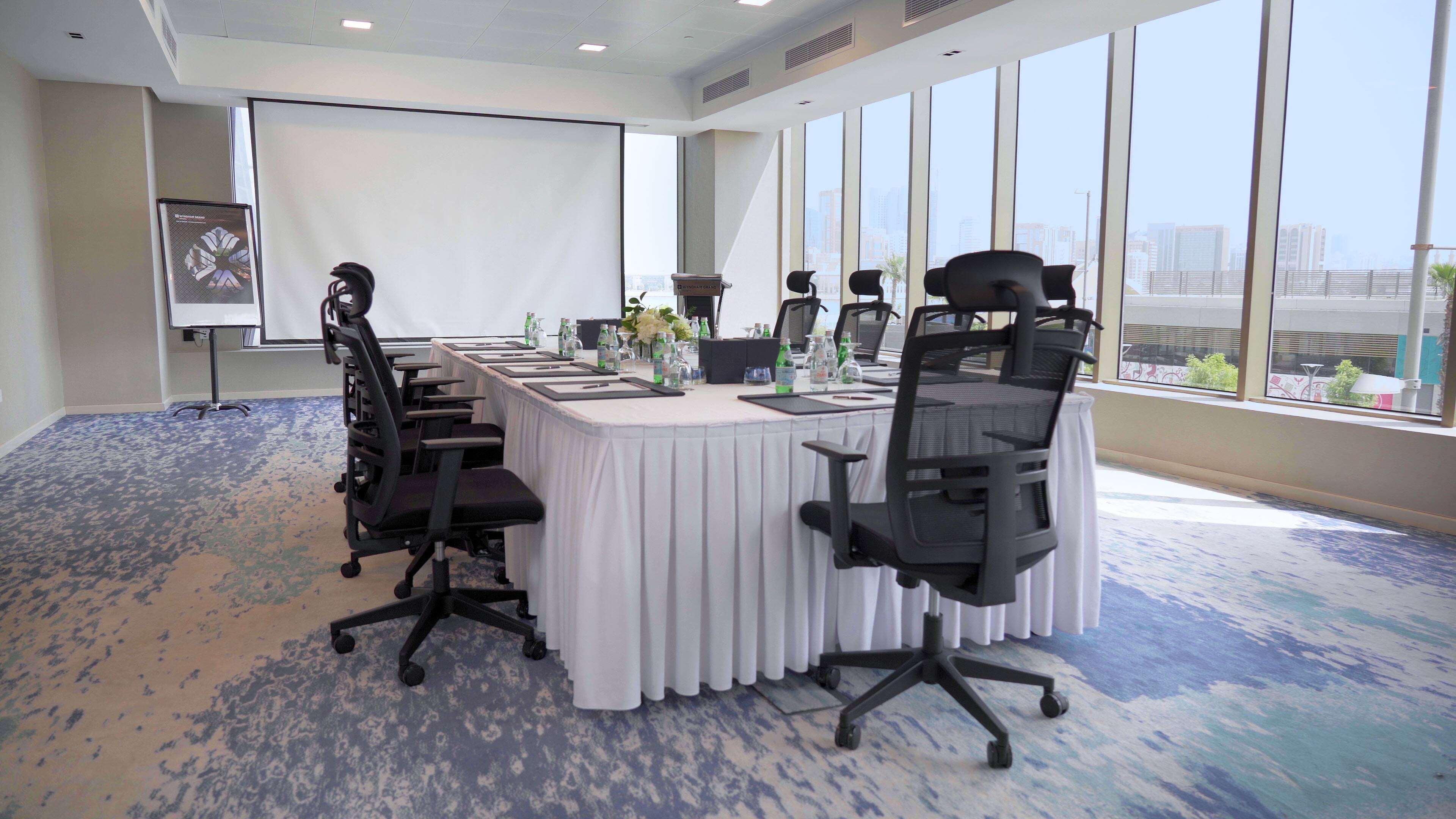 Wyndham Grand Manama Hotel Exterior photo The photo shows a modern conference room setup. There is a large rectangular table covered with a white tablecloth at the center. Around the table, there are several ergonomic office chairs. The table is neatly arranged with notepads, pens, and water