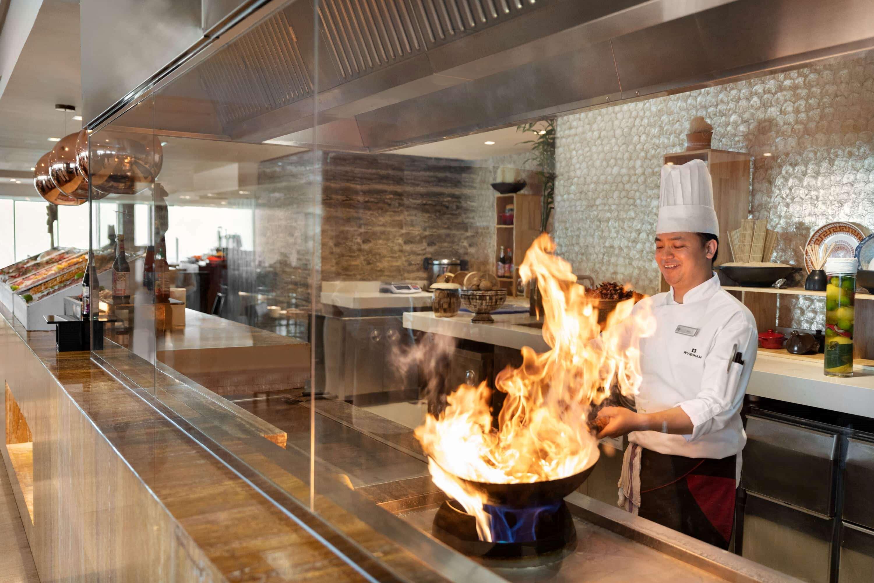 Wyndham Grand Manama Hotel Exterior photo The photo shows a chef in a professional kitchen engaged in cooking. The chef is wearing a traditional white culinary uniform and a tall white chef's hat. They are standing in front of a large wok that is producing a significant flame, indicating a h