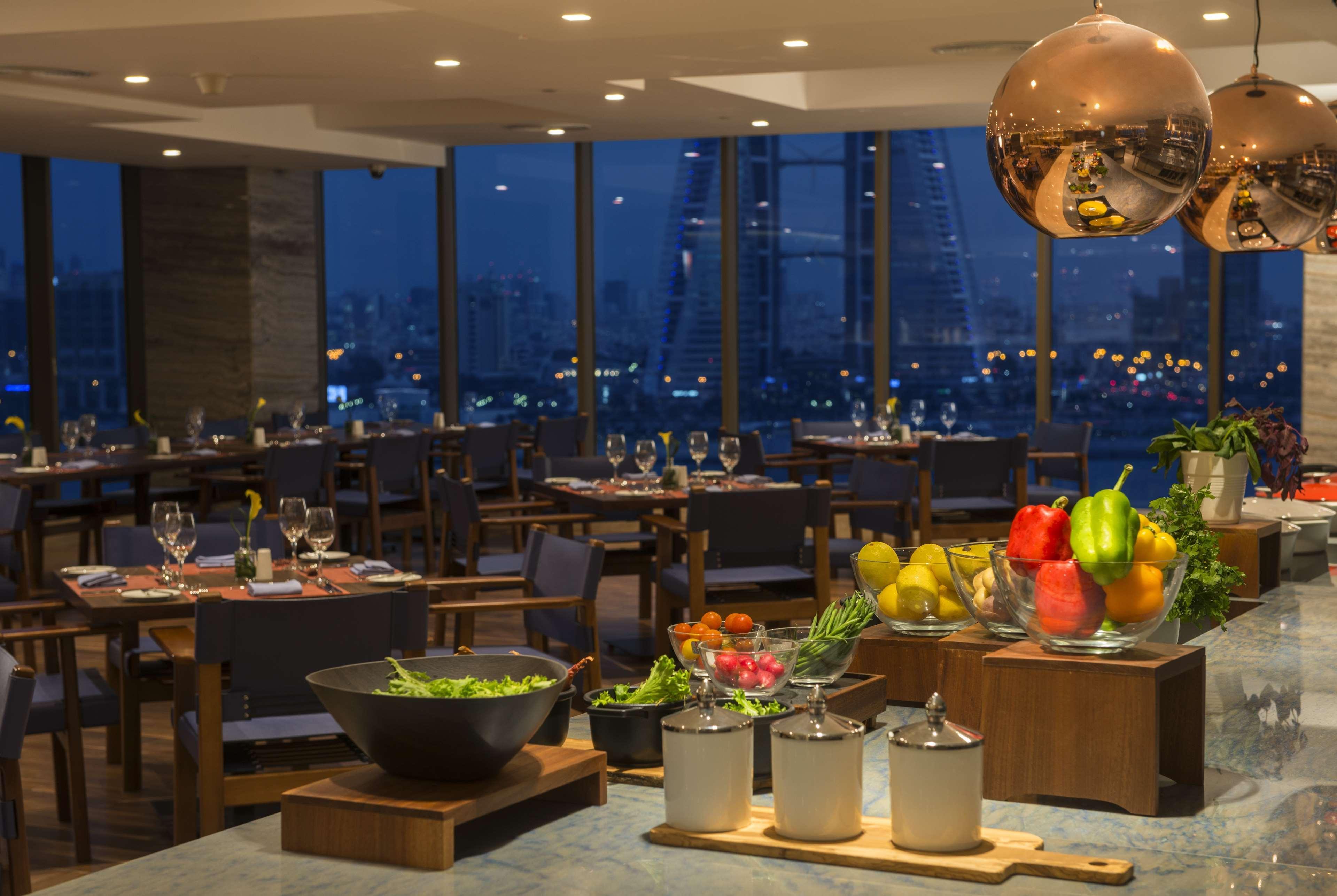 Wyndham Grand Manama Hotel Exterior photo The photo shows a stylish restaurant interior illuminated by soft lighting. There are several dining tables set for meals, with elegant dinnerware and glasses. The large windows reveal a nighttime cityscape outside, showcasing lights from buildings a