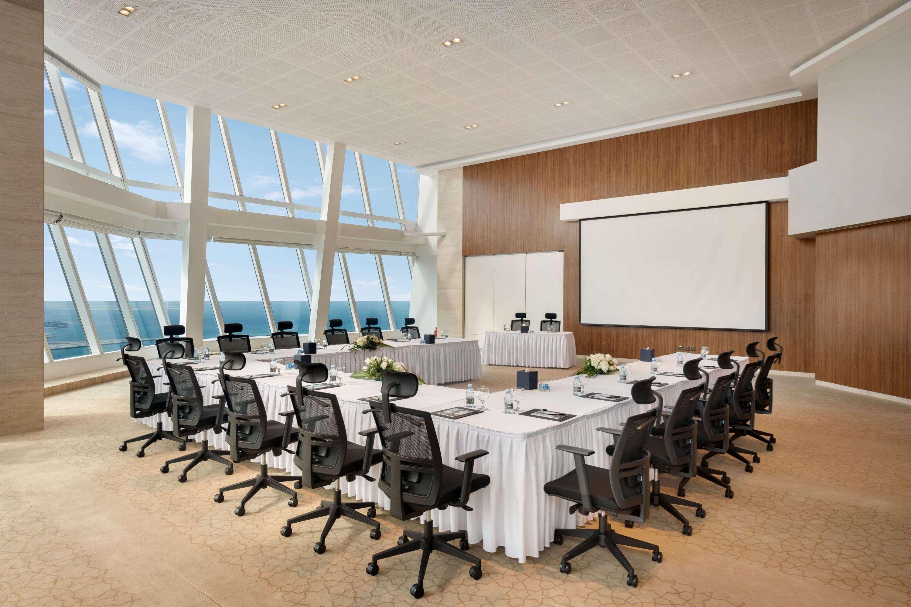 Wyndham Grand Manama Hotel Exterior photo The photo shows a modern conference room designed for meetings. It features a large oval table surrounded by black ergonomic chairs. The table is set with white tablecloths and blue notepads, creating a professional atmosphere. There are flowers in t