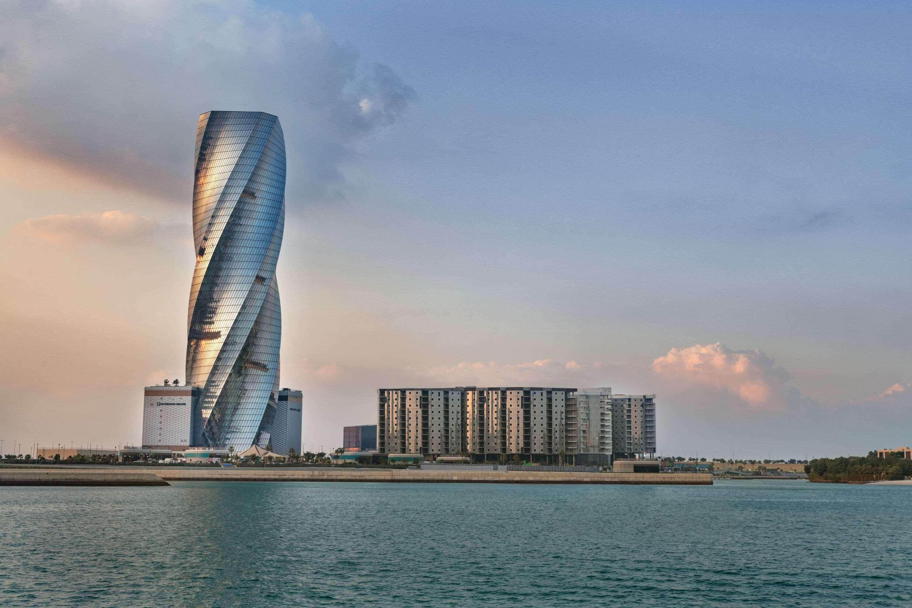 Wyndham Grand Manama Hotel Exterior photo The photo shows a modern architectural structure, likely a skyscraper, characterized by its unique twisting design and reflective glass surface. It is situated next to a body of water, which adds to the scenic quality of the image. In the foreground,