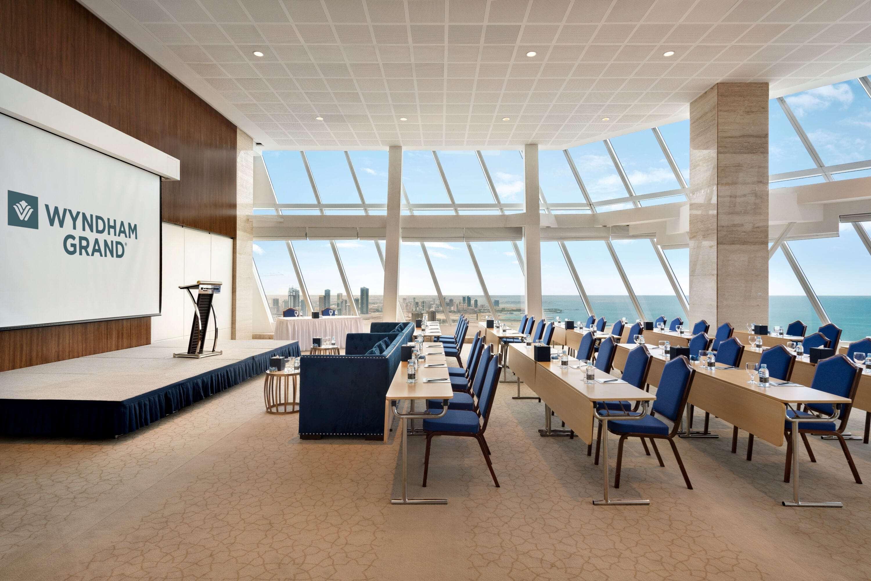 Wyndham Grand Manama Hotel Exterior photo The photo depicts a spacious conference room designed for events or meetings. It features a stage or podium at the front with a large screen displaying the name "Wyndham Grand." The room is filled with rows of seating, each with blue chairs arranged 