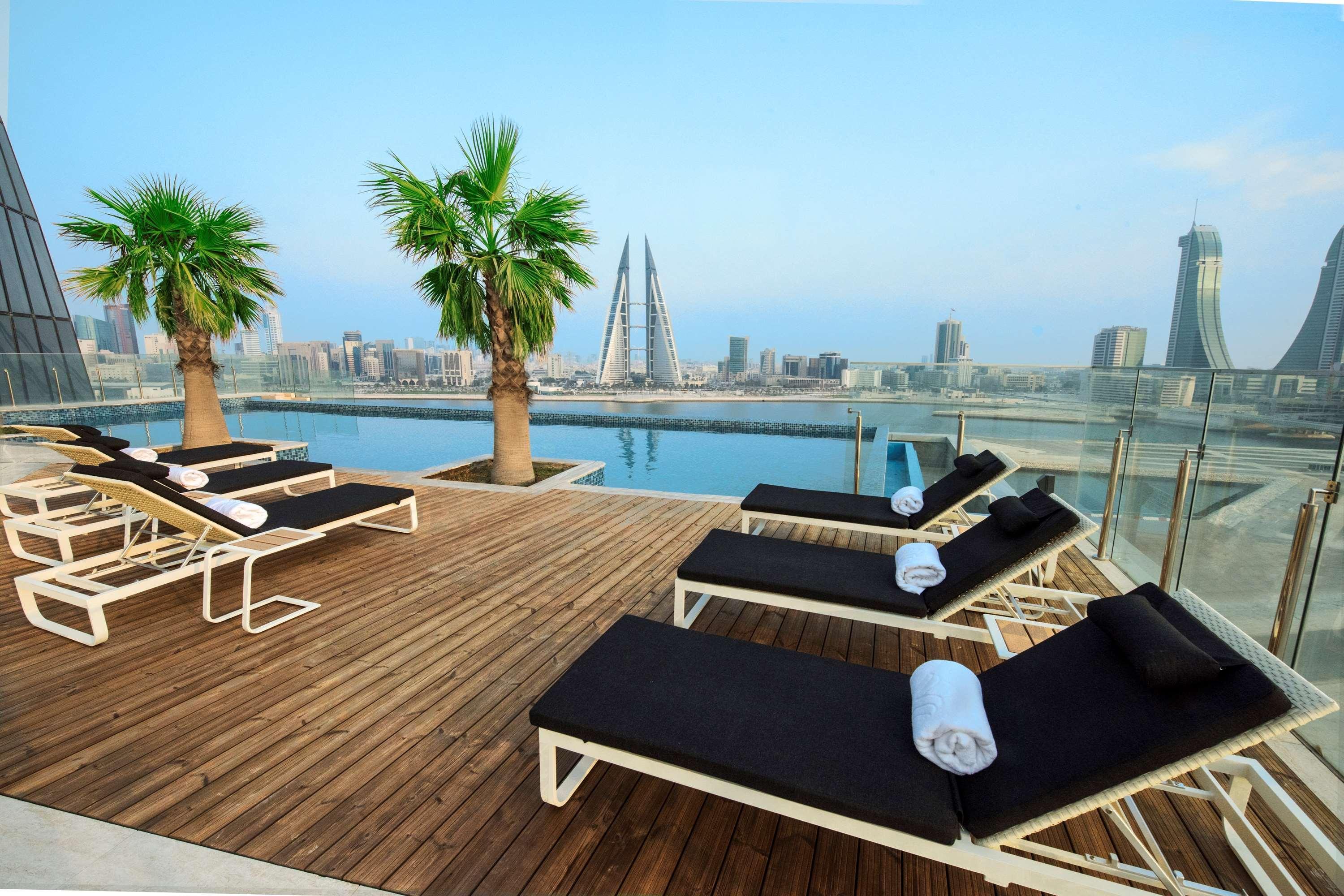 Wyndham Grand Manama Hotel Exterior photo The photo showcases a rooftop pool area with several lounge chairs arranged neatly on a wooden deck. There are palm trees placed next to the chairs, adding a tropical vibe. In the background, a city skyline is visible, featuring tall buildings, inclu
