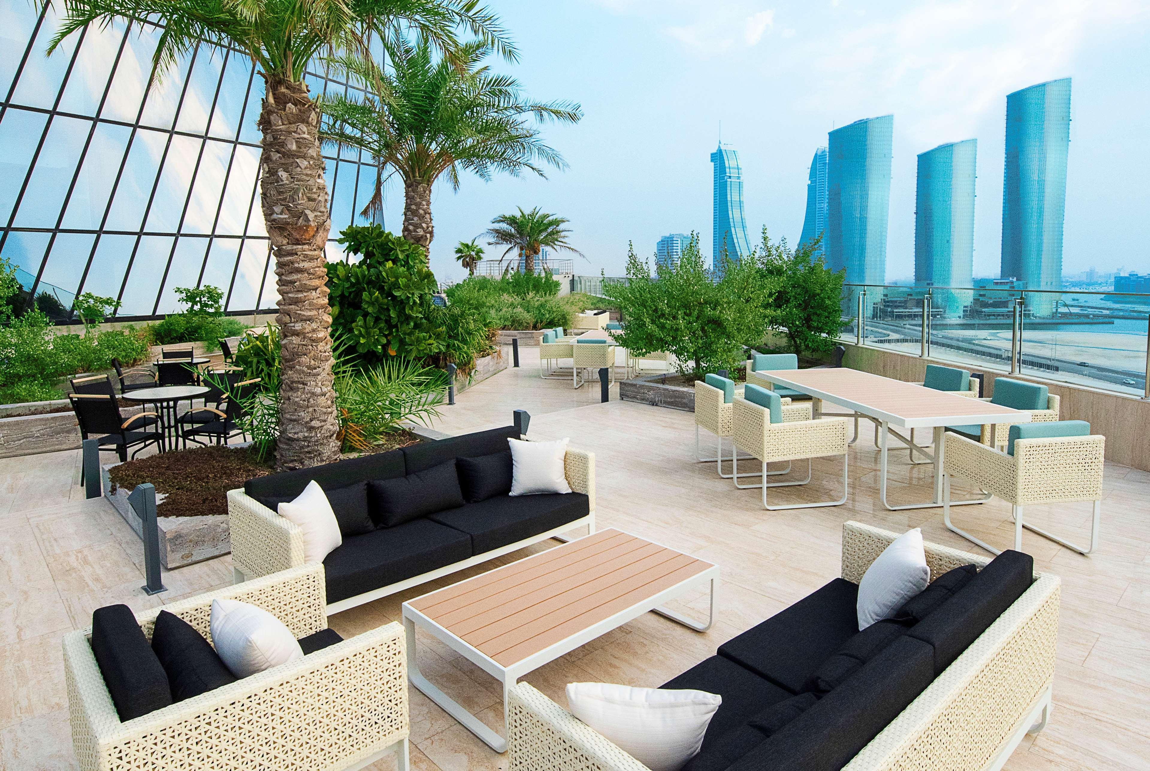 Wyndham Grand Manama Hotel Exterior photo The photo showcases a stylish rooftop lounge area featuring modern furniture. There are several seating arrangements, including black sofas with white cushions and outdoor tables surrounded by greenery. Tall palm trees add a tropical feel, while the 