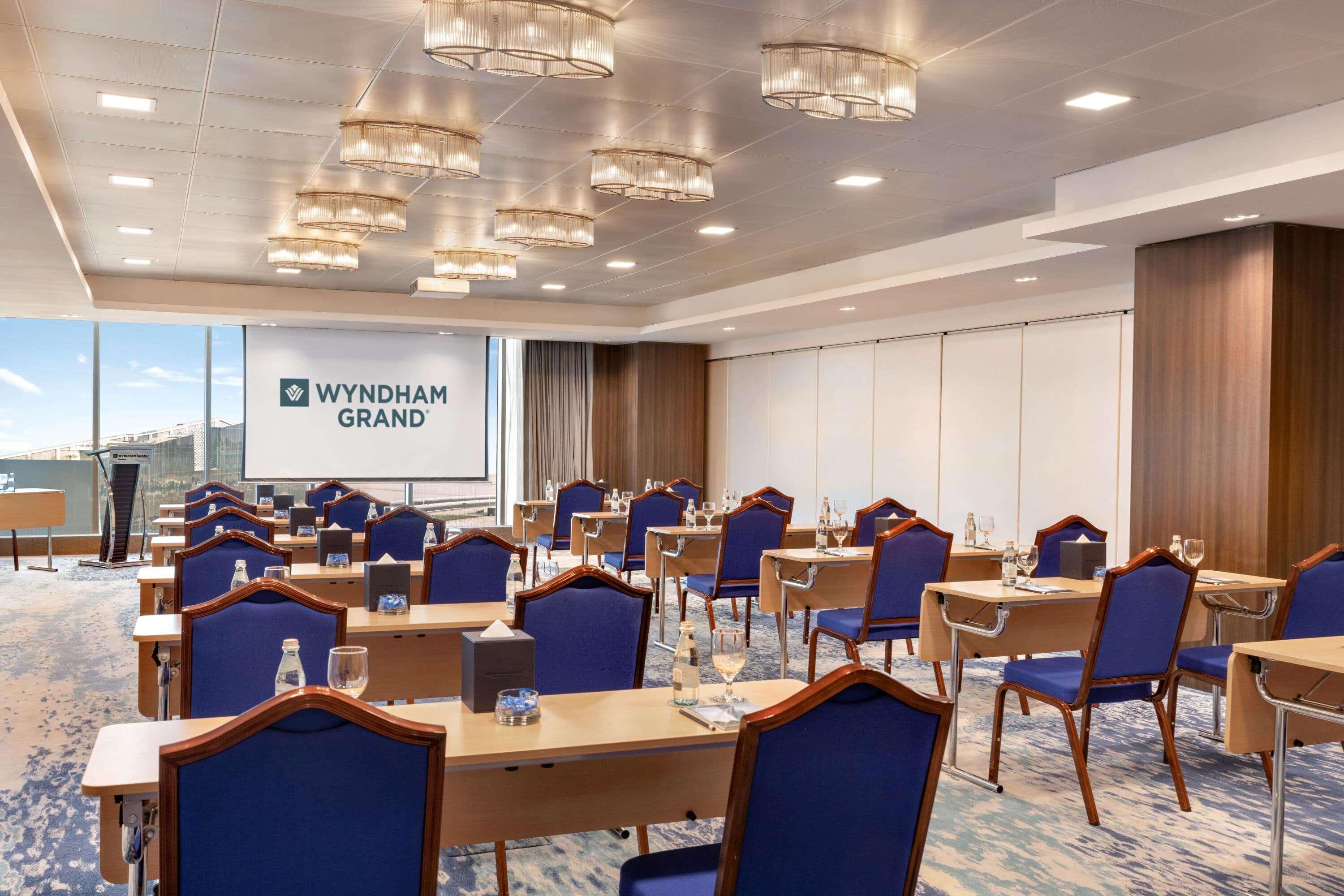 Wyndham Grand Manama Hotel Exterior photo The photo shows a conference room set up for a meeting or event. The room features several rows of tables arranged for attendees, each equipped with chairs that have blue upholstery. In the front, there is a large screen displaying the "Wyndham Grand
