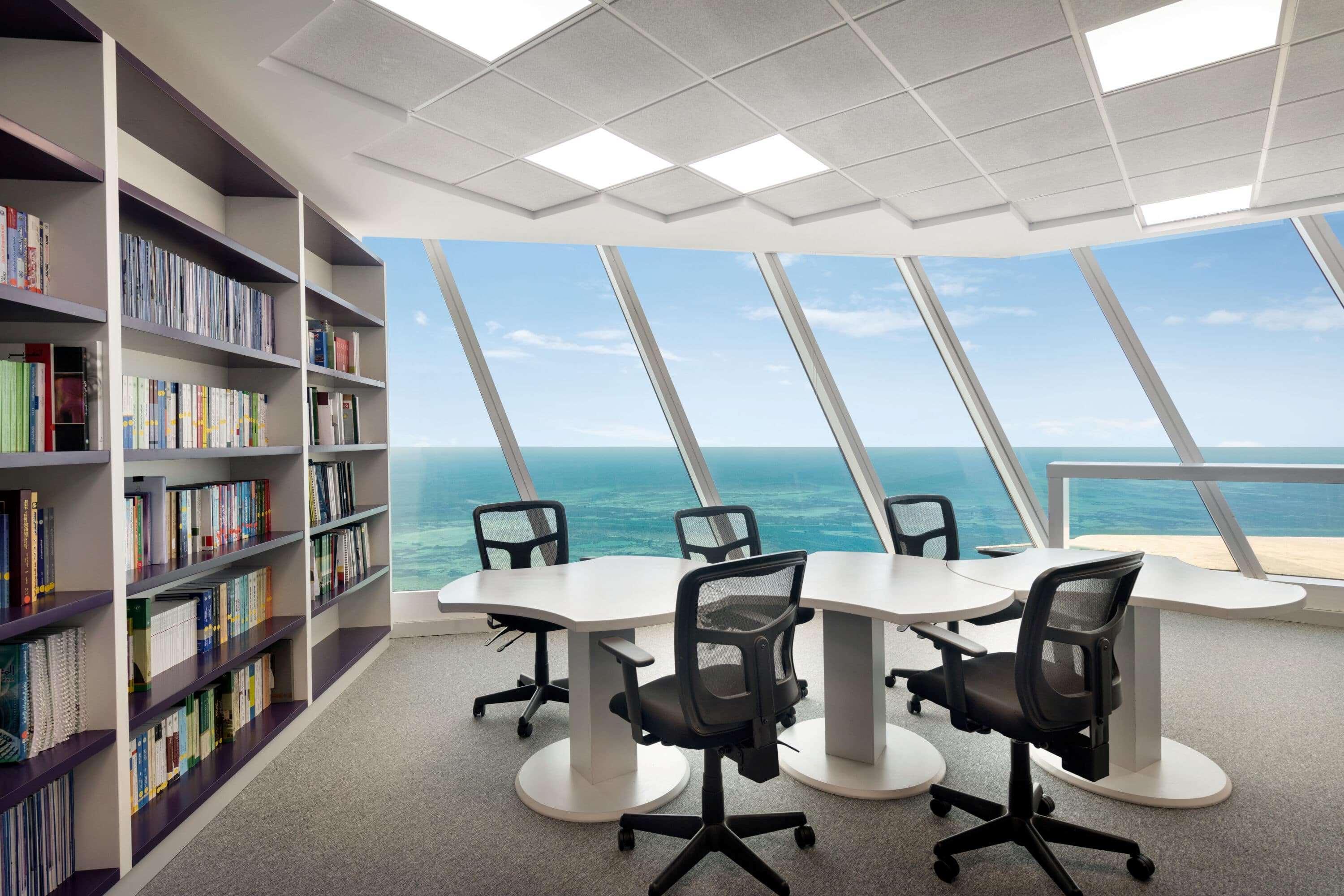 Wyndham Grand Manama Hotel Exterior photo The photo shows a modern office conference room featuring a large window with a panoramic view of the ocean. Inside, there is a round table surrounded by several black ergonomic chairs. To the left, there is a bookshelf filled with various books. The