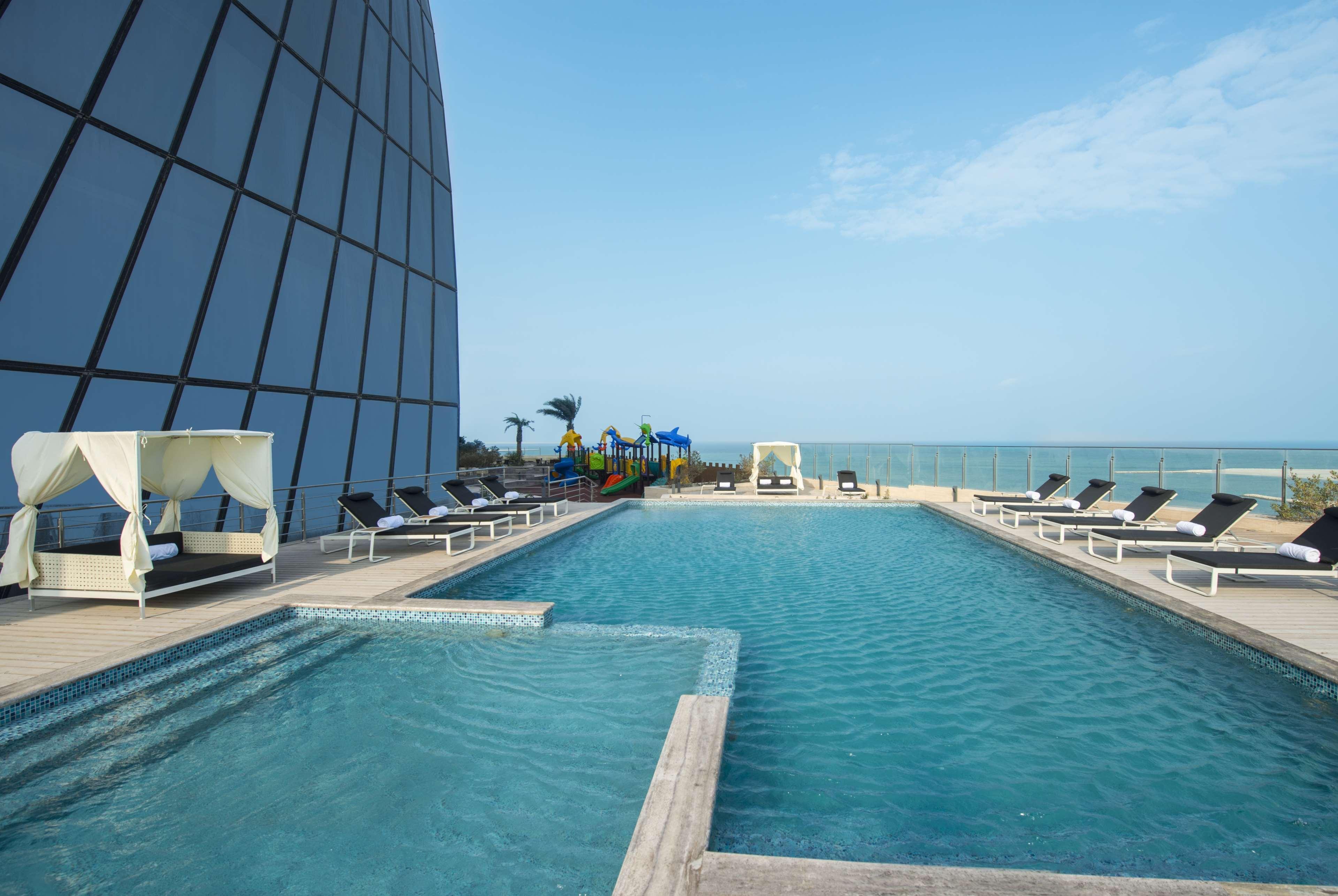 Wyndham Grand Manama Hotel Exterior photo The photo shows a rooftop pool area with a sleek, modern building in the background. The pool is large and has a few lounge chairs arranged around it. To the side, there is a smaller jacuzzi-like pool. In the distance, you can see a playground and tr