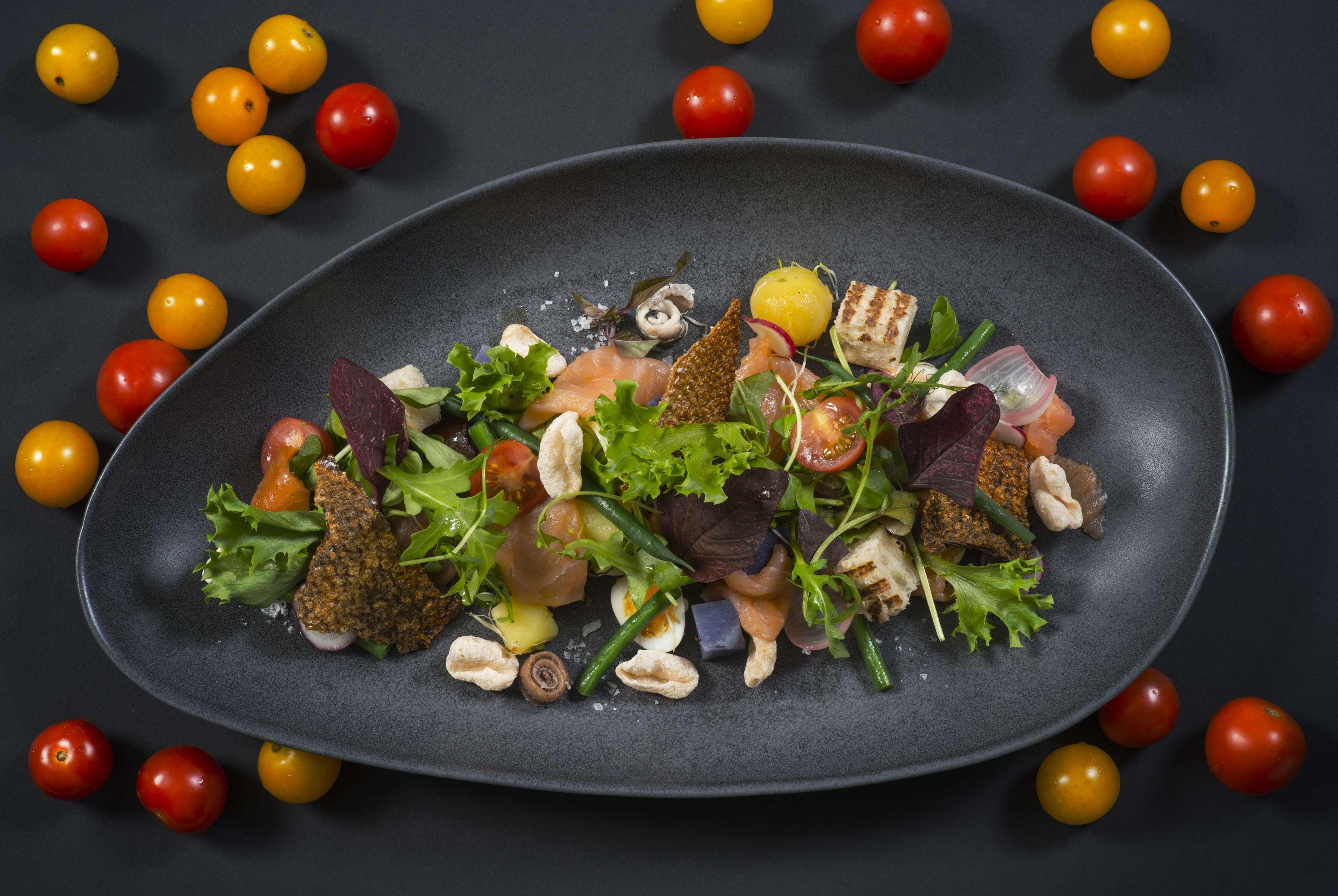 Wyndham Grand Manama Hotel Exterior photo The photo features an elegant dish presented on a dark oval plate. The salad includes a variety of fresh greens, such as leafy greens and herbs, alongside colorful cherry tomatoes in red and yellow hues. There are also pieces of seafood or fish inter