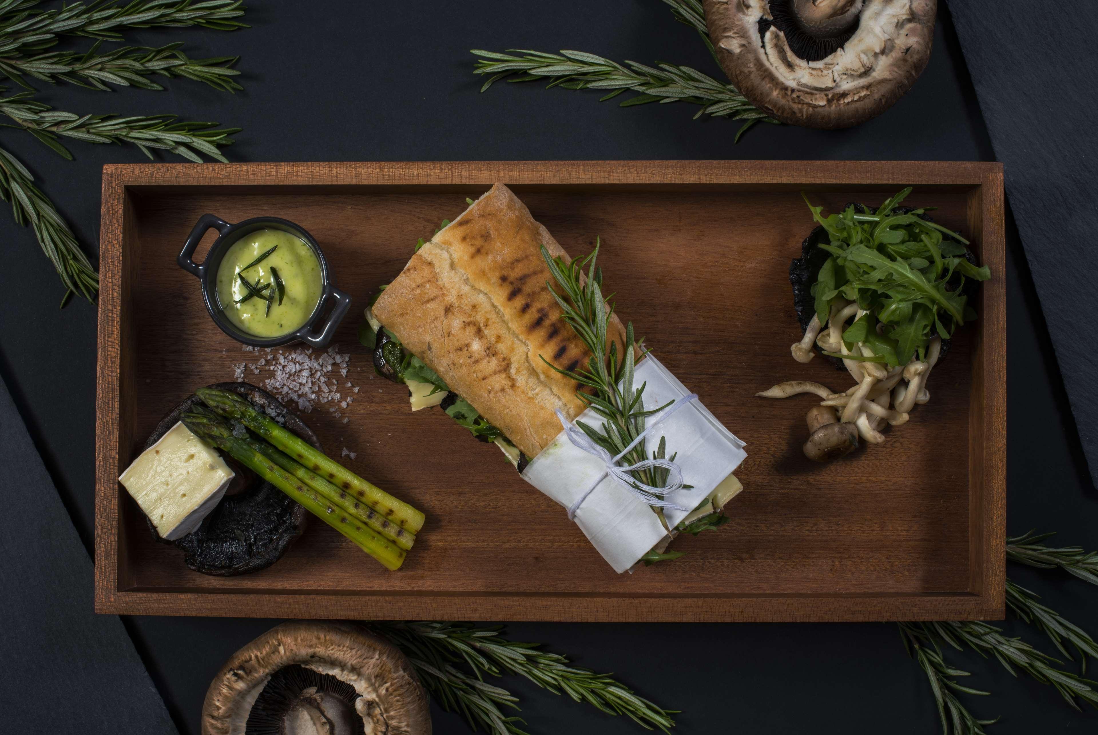 Wyndham Grand Manama Hotel Exterior photo The photo features a beautifully arranged meal on a wooden tray. It includes a large sandwich, which appears to be wrapped and garnished with herbs. On the side, there are several small portions of food: grilled asparagus, a piece of cheese on a blac