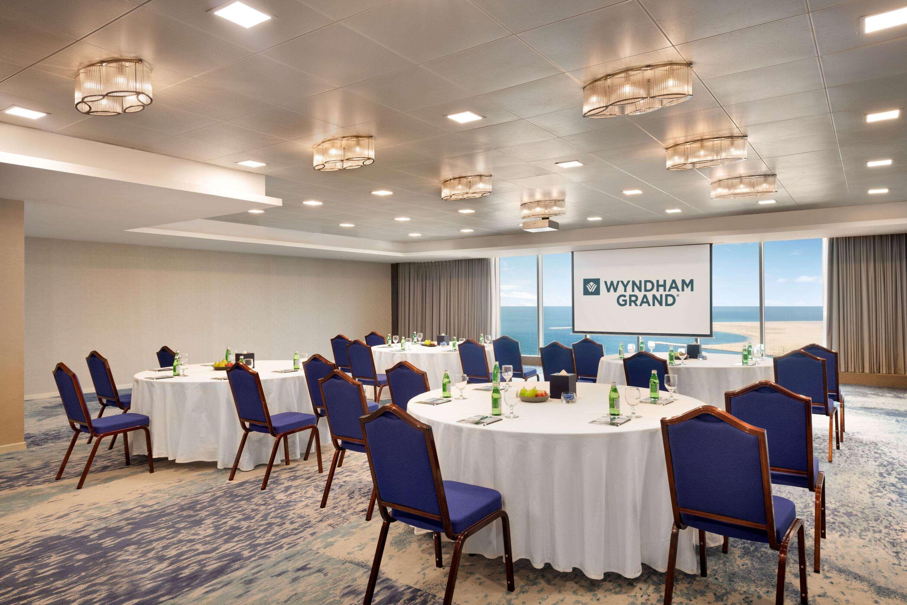 Wyndham Grand Manama Hotel Exterior photo The photo shows a conference or event room set up for a meeting at the Wyndham Grand hotel. It features several round tables, each with white tablecloths and blue chairs around them. On one wall, there is a large presentation screen displaying the Wy