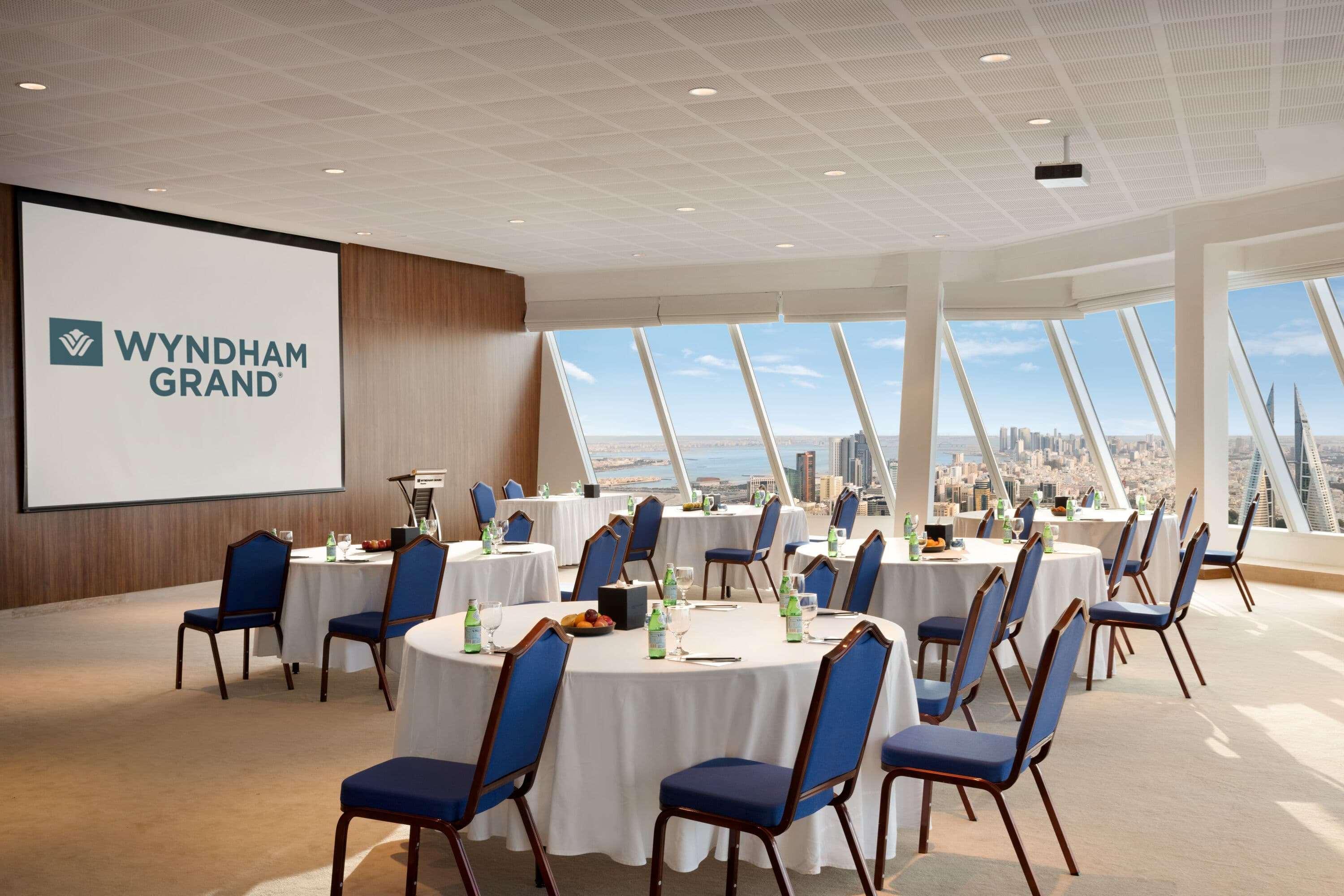 Wyndham Grand Manama Hotel Exterior photo The image shows a modern conference room or meeting space that appears to be part of the Wyndham Grand hotel. The room features several round tables covered with white tablecloths, each accompanied by blue chairs. There are green bottles of water on 