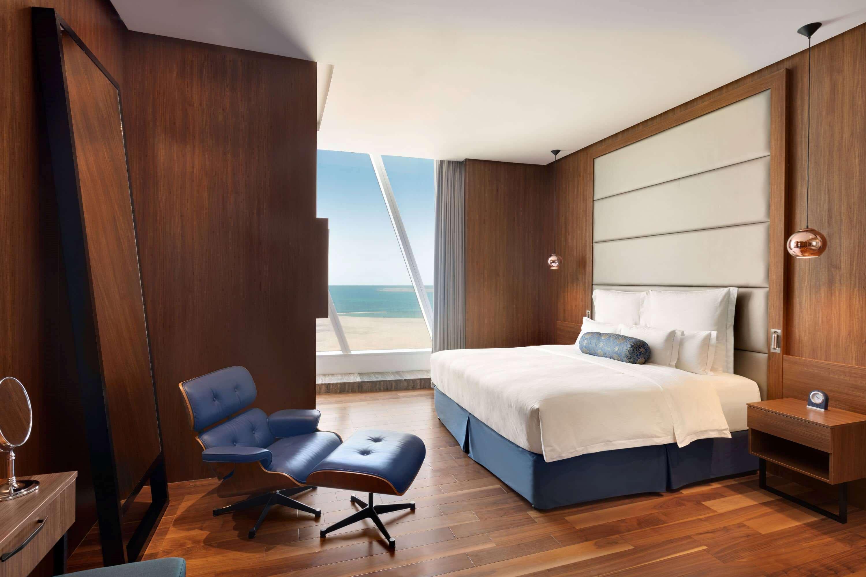 Wyndham Grand Manama Hotel Exterior photo The photo depicts a modern hotel room with a spacious and stylish design. It features a large bed with white bedding situated against a wood-paneled wall. To the side, there is a comfortable blue armchair and a small side table. The room has large wi