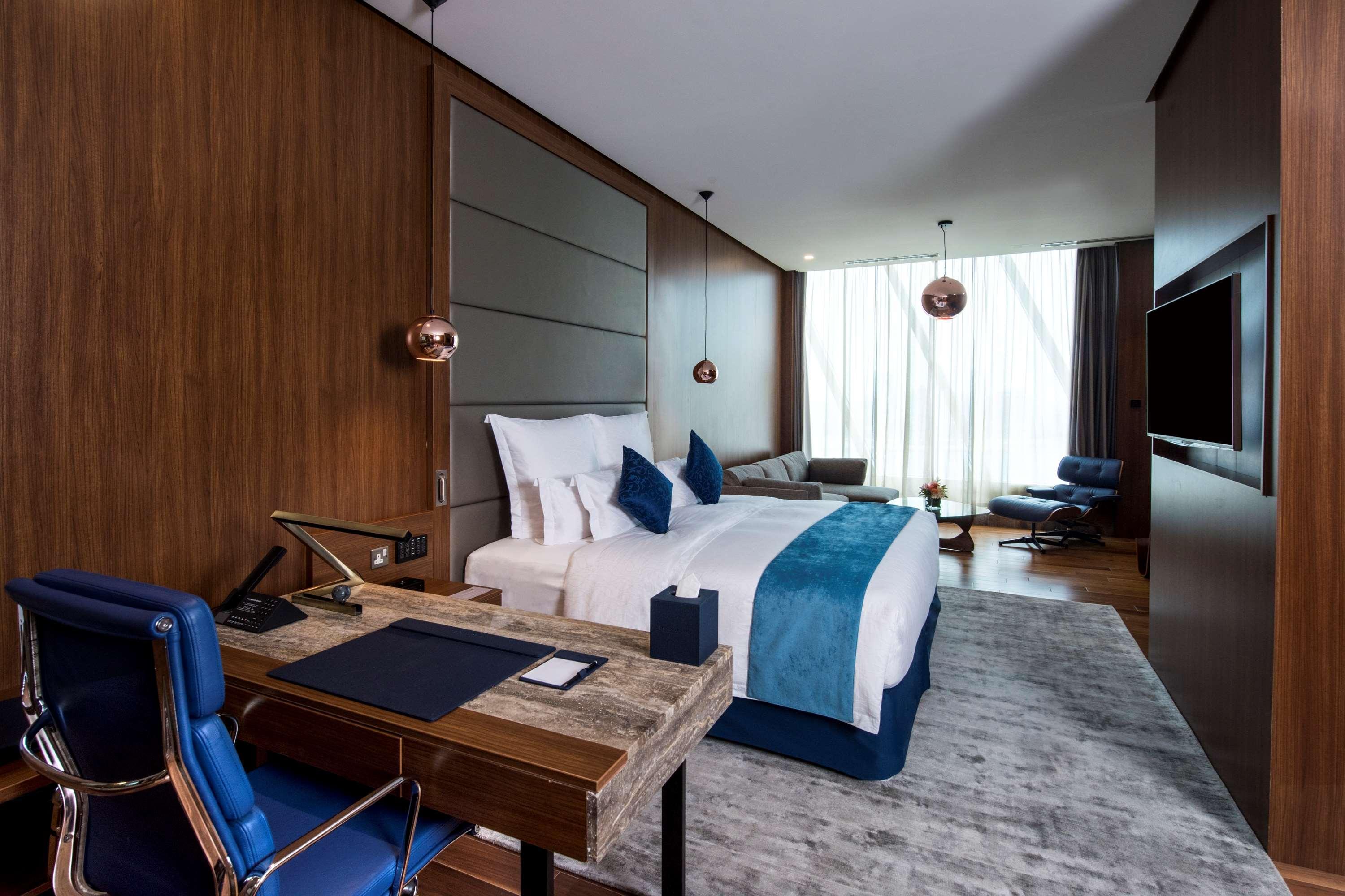 Wyndham Grand Manama Hotel Exterior photo The photo shows a modern hotel room with a sleek and stylish design. It features a large bed with white linens and decorative blue pillows. The room has wooden paneling on the walls, creating a warm and inviting atmosphere. A comfortable seating area