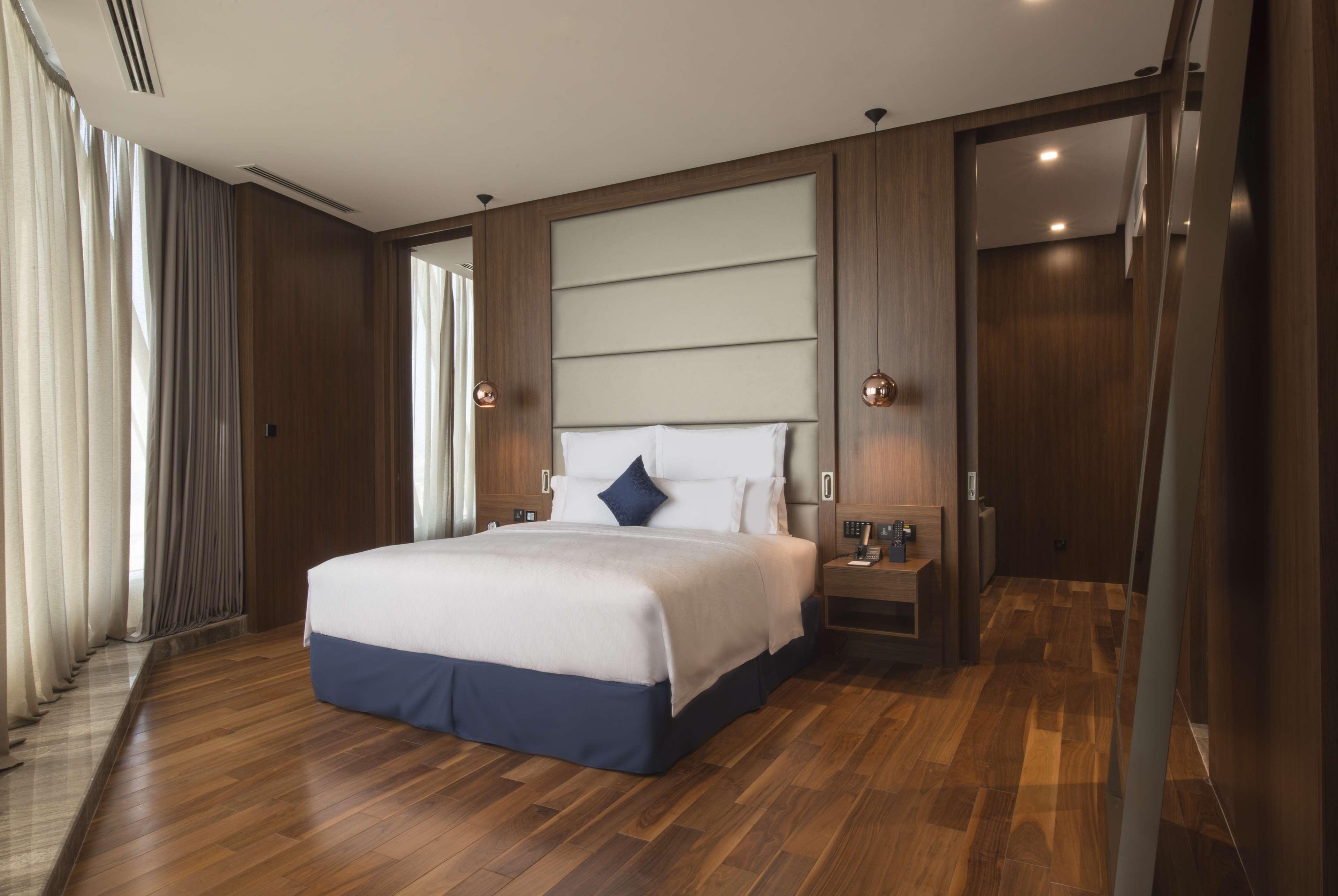 Wyndham Grand Manama Hotel Exterior photo The photo shows a modern hotel room. It features a queen-sized bed with crisp white linens and a decorative blue accent pillow. The walls have a wood finish, creating a warm and inviting atmosphere. Large windows with sheer curtains allow natural lig