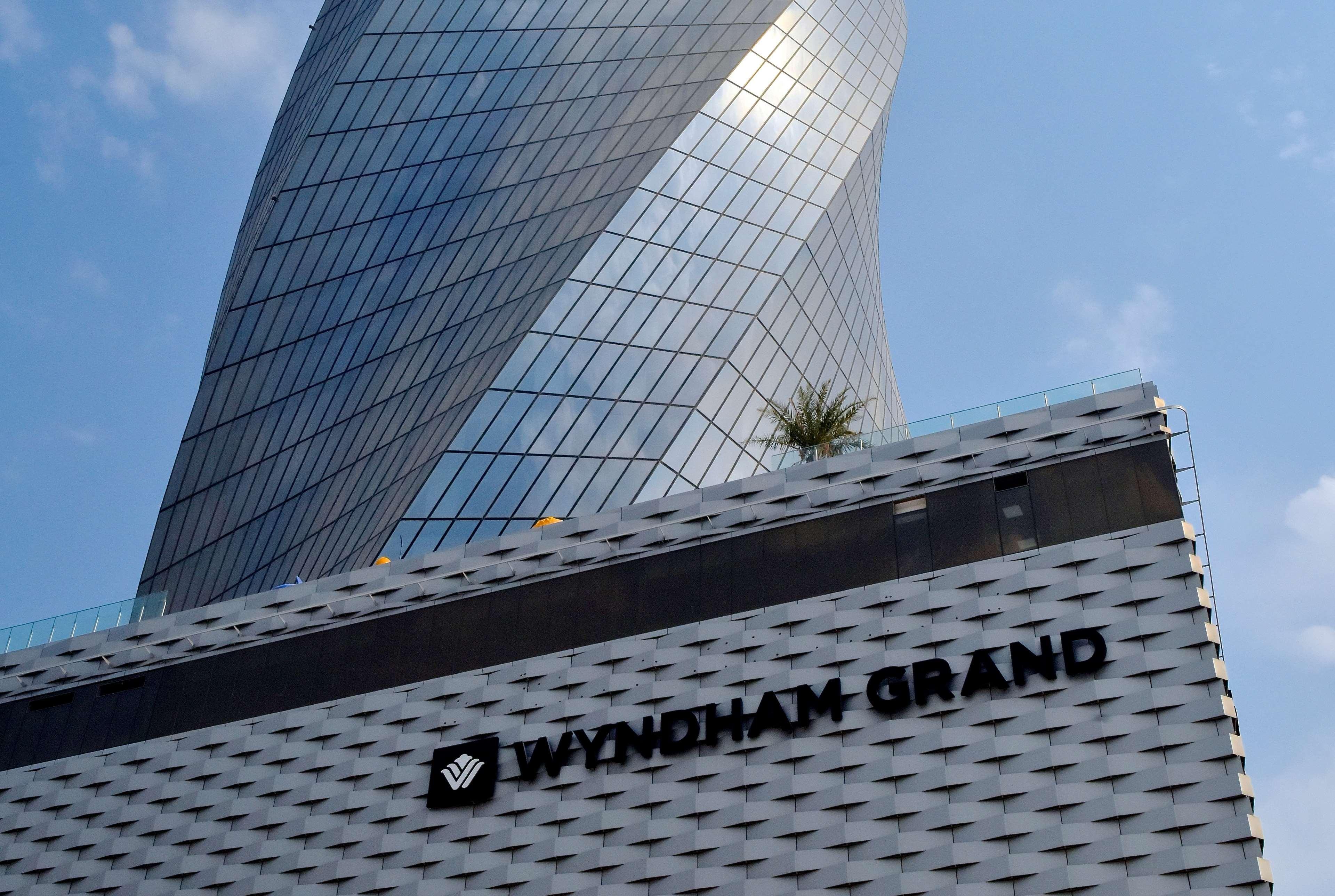 Wyndham Grand Manama Hotel Exterior photo The photo shows a modern, tall building with a distinctive curved and angular design. The structure features reflective glass windows that create a shiny appearance. At the bottom of the building, there is a sign displaying the name "Wyndham Grand" i